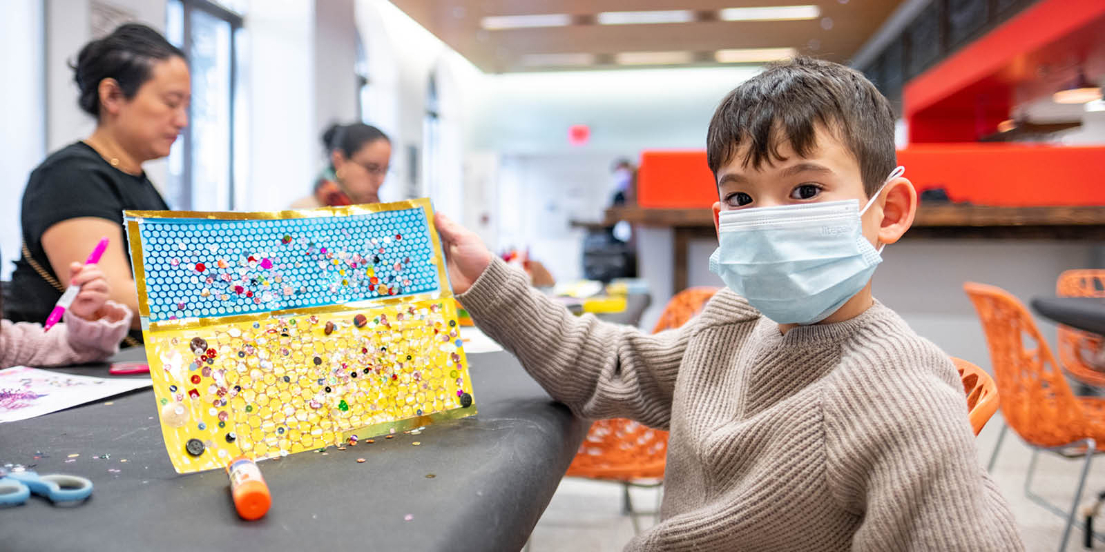 A boy wearing a face mask holding an arts and crafts project made from yellow and blue paper with small round buttons attached